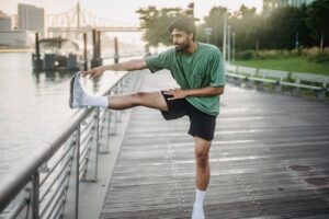 Whole body calisthenics workout warm up, man doing a standing hamstring stretch with one leg on a waterfront railing.